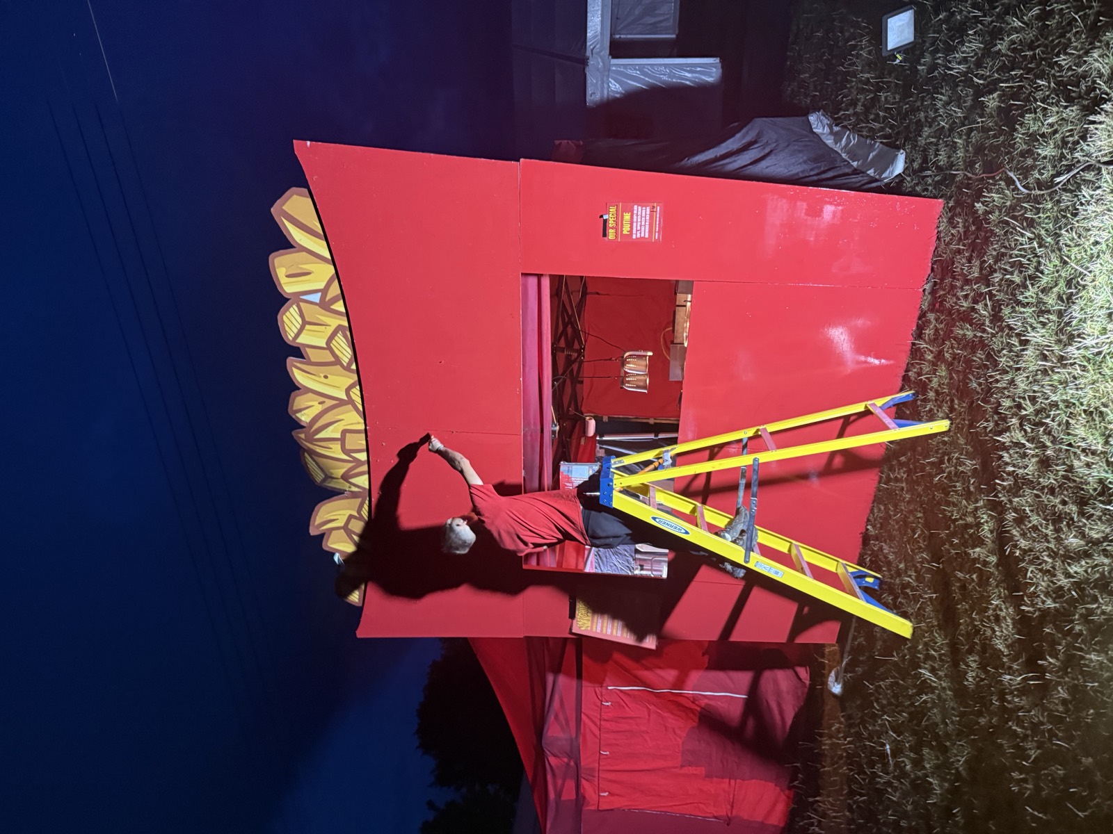 Building a giant chip-box food stall in a field at night, someone up a ladder